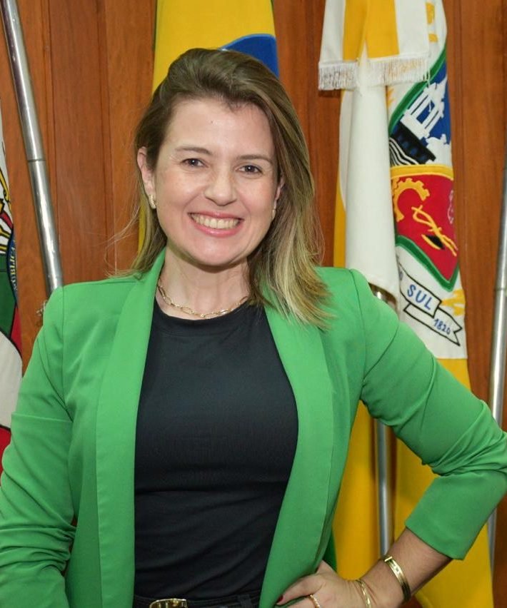Ana Paula Melo sorri, de blazer verde, diante das bandeiras da Câmara de Vereadores