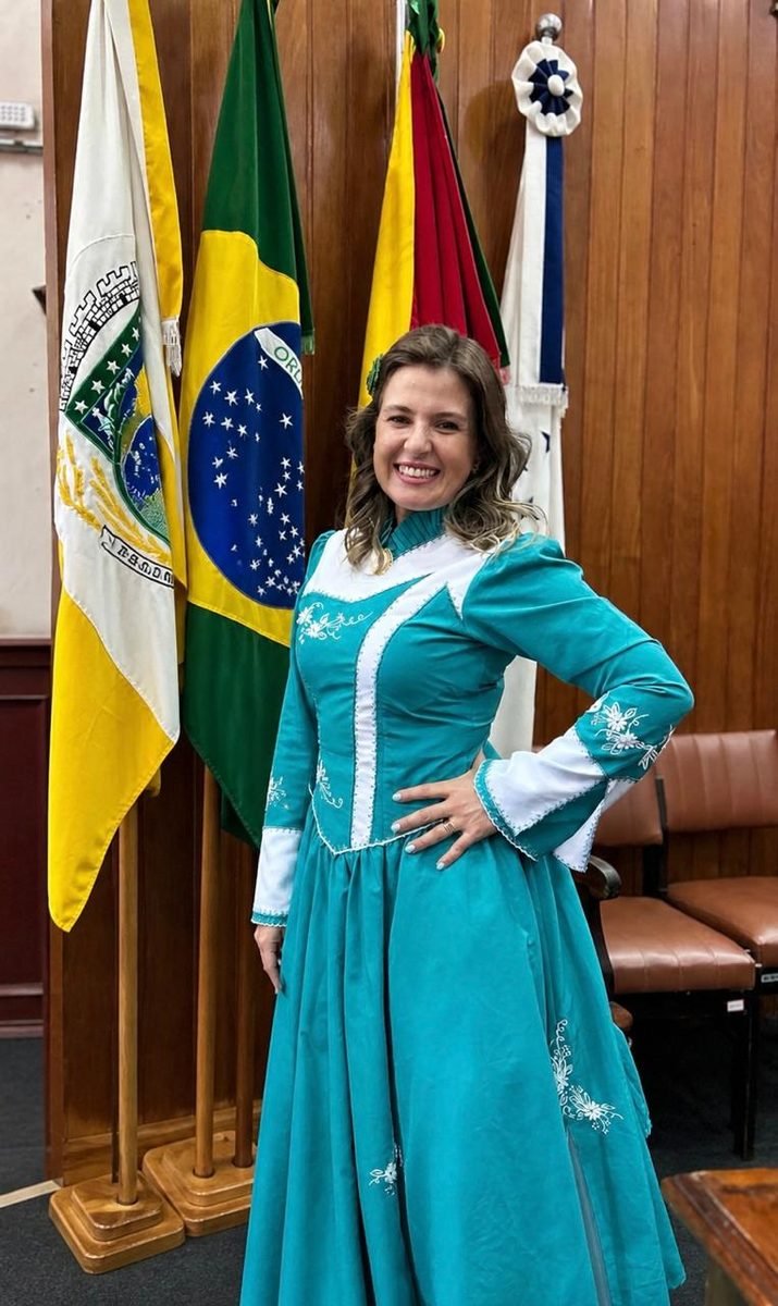 Ana Paula Melo com vestido de prenda azul-turquesa, diante das bandeiras no plenário